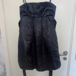 Svart klänning med smala axelband - En elegant svart klänning med smala axelband som går att ta bort och subtilt mönster. Klänningen har en rak passform och är knälång. Den är tillverkad av ett glansigt material som ger en lyxig känsla.