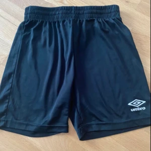 Svarta shorts från Umbro - Ett par svarta shorts från Umbro i storlek 164 (13-14 år). Shortsen har en elastisk midja och är tillverkade i ett lätt och bekvämt material.
