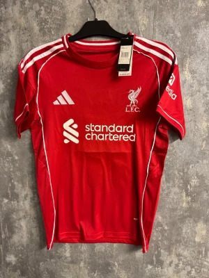 Liverpool M. Salah matchtröja röd - S - Liverpool FC matchtröja i rött med vita detaljer, Adidas-logga och Standard Chartered-tryck på bröstet. På ryggen står M. Salah och nummer 11. Tröjan har klassiska vita ränder på axlarna och är tillverkad i lätt polyester för bästa komfort.