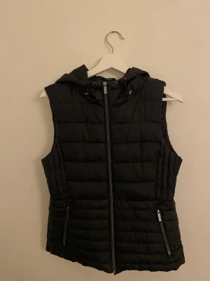 Svart dunväst med huva från Zara - stl M - Snygg svart vadderad väst från Zara i bra skick. Lätt men varm, perfekt som lager-på-lager under vår/höst eller över hoodie och tröja. Dragkedja framtill, fickor med dragkedja och huva. 