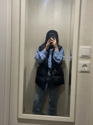 Svart dunväst med snörning - Säljer en svart, lång och puffig dunväst med snörning i midjan. Västen har en hög krage och stängs med dragkedja framtill. Perfekt att slänga över en hoodie eller skjorta för en chill och trendig look.