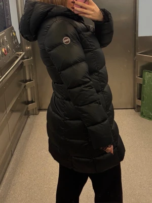 Svart colmar lång pufferjacka med huva - Säljer en Colmar svart, lång pufferjacka med huva och dragkedja framtill. Perfekt för kalla dagar när du vill hålla dig varm. Köpt för 6000kr. Inga defekter. 