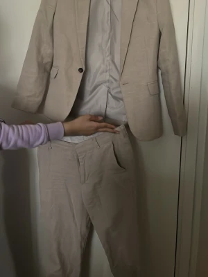 Beige kostym från H&M, strl 146 - Säljer en stilren beige kostym från H&M i storlek 146. Setet består av en kavaj med knappstängning och två fickor samt matchande kostymbyxor med fickor och hällor för bälte. Materialet är linneblandning med slät insida i syntet. Perfekt för dig som gillar klassisk look.