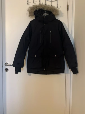 Svart dunjacka med fuskpäls från Everest - Svart dunjacka från Everest med fuskpäls på huvan, två stora fickor med knappar och två bröstfickor med dragkedja. Jackan har långa ärmar med tumhål och quiltad insida för extra värme. Perfekt för kalla vinterdagar.