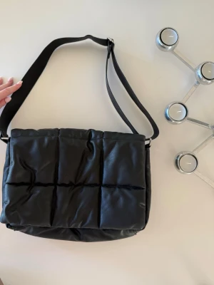 Svart quiltad axelväska från Monki - Quiltad svart axelväska från Monki i mjukt skinnimitation. Väskan har ett rutmönstrat yttre, justerbar axelrem och magnetknäppning. Insidan har ett fack med dragkedja för småsaker. Perfekt storlek för laptop, böcker eller vardagsprylar.