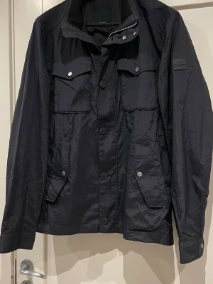 Mörkblå fieldjacket från J.Lindeberg - Mörkblå fieldjacket från J.Lindeberg Jackan har flera fickor med knappar, klassisk krage och snygga detaljer som logopatch på ärmen. Materialet är lätt och känns sportigt, perfekt för dig som gillar stilren design och funktion. Jackan är i XL men sitter mer som en L