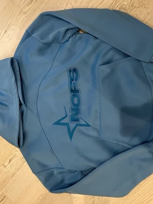 Säljer blå NOFS hoodie med huva och logga. Mjuk, skön och i bra skick. - Jag säljer en blå hoodie från NOFS. Den har huva och NOFS-logga på framsidan. Tröjan är mjuk och skön att ha på sig. Den är i bra skick utan hål eller fläckar och passar bra till vardag och fritid.