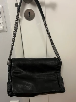 Svart axelväska med kedja - Säljer en svart axelväska i skinn med rejäl silverfärgad kedja som axelrem. Väskan har en mjuk, lite slouchy form och är enkel men ändå cool. Väskan är rymlig och du får plats med allt det nödvänliga, i väskan finns det även tre olika fack så att du kan hålla koll på dina prylar. Kedjan ger en edgy vibe och väskan funkar till många olika stilar.