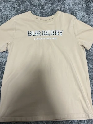 Beige Burberry t-shirt med logga - Snygg beige t-shirt från Burberry med klassisk logga och texten 'London England' på bröstet. T-shirten har rund hals och korta ärmar, perfekt för en clean och stilren look. Tillverkad i mjuk bomull för skön känsla.