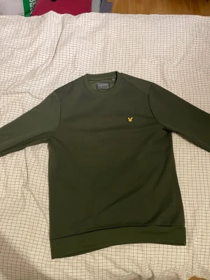 Mörkgrön sweatshirt från Lyle & Scott - Säljer en stilren mörkgrön sweatshirt från Lyle & Scott med klassisk rund hals och långärmad design. Tröjan har en gul broderad logotyp på bröstet och ribbade muddar vid ärmslut och nederkant. Perfekt för dig som gillar enkel och snygg streetstil. 