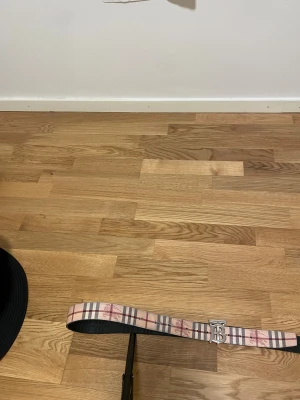 Burberry bälte - Säljer ett snyggt Burberry-bälte i skinn med klassiskt rutig design i beige, röd och svart. Bältet har en silverfärgad metallspänne med Burberrys ikoniska TB-logga. Perfekt för att lyfta din outfit med en lyxig touch.
