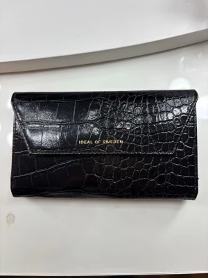 Svart plånboksväska Ideal of Sweden - Snygg svart plånboksväska från Ideal of Sweden med croco-mönster i skinnimitation. Väskan har plats för mobil och kort, och stängs med magnet. Passar iPhone 14/15