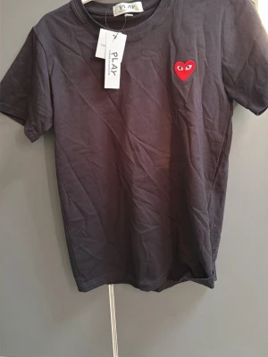 Svart t-shirt Comme des Garçons Play - Svart t-shirt från Comme des Garçons Play med klassisk rund hals och korta ärmar. På bröstet sitter det ikoniska röda hjärtat med ögon, broderat. Materialet är mjuk bomull och passformen är normal. Perfekt för dig som gillar streetwear och stilrena detaljer.