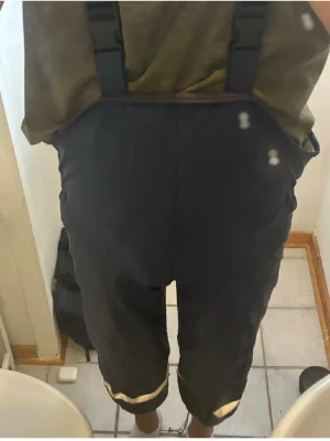 Svarta täckbyxor med hängslen - Säljer ett par svarta täckbyxor med justerbara hängslen och reflexdetaljer vid bensluten. Byxorna är tillverkade i ett slitstarkt syntetmaterial och har en loose passform som ger bra rörelsefrihet. Perfekta för kalla dagar och vinteraktiviteter.