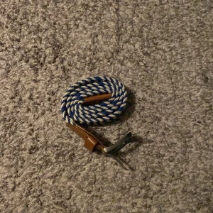 Randigt blåvitt bälte med skinn - Säljer ett snyggt flätat bälte med randigt mönster i blått och vitt. Bältet har detaljer i brunt skinn vid ändarna och ett klassiskt metallspänne. Perfekt accessoar för att lyfta din outfit med lite färg och stil.