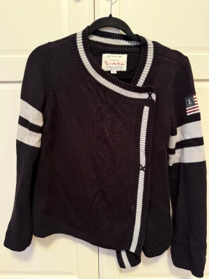 Svart stickad kofta från Bondelid - Svart stickad kofta från Bondelid med gråa ränder på ärmarna och runt kragen. Snygga detaljer med ribbad kant och mönsterstickning framtill och baktill. Långärmad och med patch på ena ärmen. 