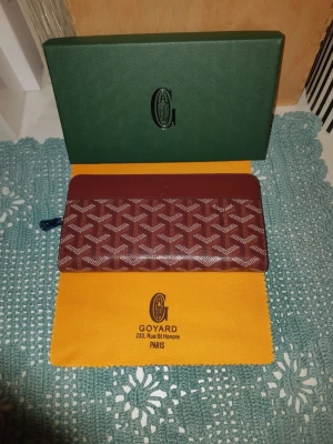 Brun Goyard plånbok med mönster - Säljer en brun plånbok från Goyard med deras ikoniska vita mönster och präglad logga. Plånboken har dragkedja runtom och flera fack inuti för kort och sedlar. Materialet är skinn och canvas, och designen är riktigt clean och stilren.