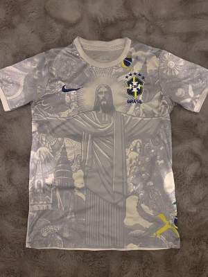 Vit Brasil Nike t-shirt med tryck - Snygg vit Brasil t-shirt från Nike med stort tryck av Kristusstatyn och detaljerade grafiska mönster på framsidan. CBF-logga och Nike swoosh i blått, samt inslag av gult och grönt. Tillverkad i lätt polyester, perfekt för fotboll eller streetstyle.