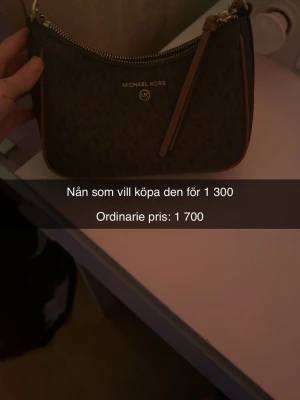 Brun handväska Michael Kors - Säljer en snygg brun handväska från Michael Kors med MK-logga och guldiga detaljer. Väskan har ett mönstrat yttre med MK-symboler, dragkedja upptill och flera fack inuti. Perfekt storlek för det viktigaste och har en stilren look.