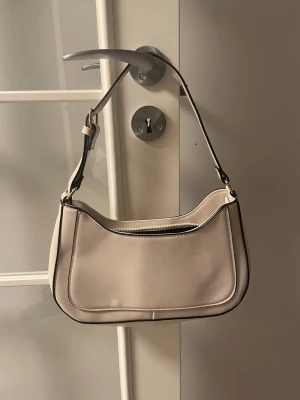 Beige axelväska med dragkedja - Snygg beige axelväska med stilren design och svarta detaljer längs kanterna. Väskan har en praktisk dragkedja upptill och ett justerbart axelband. Perfekt storlek för det viktigaste och passar till många olika outfits.