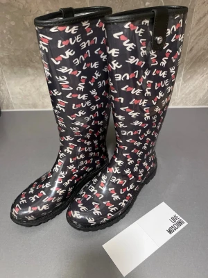 Love Moschino svarta stövlar med print - Svarta gummistövlar från Love Moschino med mönster i vitt och rött över hela skon.  Stövlarna har rund tå, platt grov sula och är gjorda i syntetmaterial som tål regn. Perfekt statement-piece med box och originalpapper. Storlek 38. Mycket bra skick, som nya! Säljs på flera sidor, först till kvarn. 