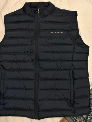 Svart dunväst från J.Lindeberg - Snygg mörkblå dunväst från J.Lindeberg med diskret logga på bröstet. Västen har quiltad design och dragkedja framtill. Perfekt att slänga på över en hoodie eller tröja när det är lite kyligt ute.