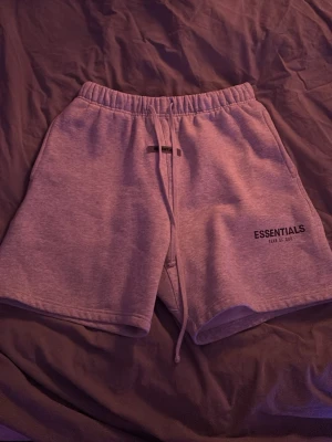 Grå Essentials Fear of God shorts - Säljer ett par grå mjukisshorts från Essentials Fear of God. De har snörning i midjan, en bakficka och tryckt logga på framsidan. Perfekt för chill dagar och har en loose passform som är riktigt skön. Materialet är mjukt och känns som bomull. Ordinarie pris ligger på 1299 och dem är knappt andvända. Hör gjärna av er vid funderingar🤩🔥