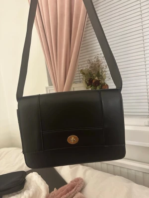 Svart axelväska med guldspänne - Stilren svart axelväska i skinnimitation med bred axelrem och elegant guldigt vridlås framtill. Väskan har en rektangulär form och slät yta utan mönster, perfekt för dig som gillar minimalistisk design. Insidan är rymlig och praktisk för dina viktigaste grejer.