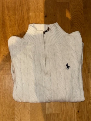 Vit stickad polotröja från Ralph Lauren - Säljer en vit stickad polotröja från Ralph Lauren med klassiskt kabelmönster och halv dragkedja framtill. Tröjan har hög krage och den ikoniska loggan broderad på bröstet. Perfekt för kyliga dagar och snygg till jeans.