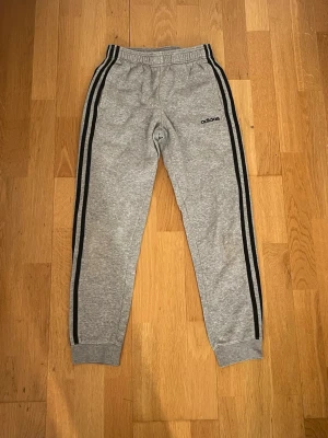 Grå Adidas mjukisbyxor med svarta ränder - Grå mjukisbyxor från Adidas med klassiska svarta ränder längs sidorna och Adidas-logga på låret. Byxorna har resår och snörning i midjan samt muddar vid bensluten. 