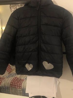 Svart pufferjacka med reflexhjärtan - Säljer en svart pufferjacka med huva och dragkedja. Jackan har flera silvriga reflexhjärtan på fram- och baksidan samt på ärmarna. Perfekt för dig som vill synas i mörkret och ändå hålla stilen. Jackan är quiltad och har en normal passform.luvan är avtagbar.