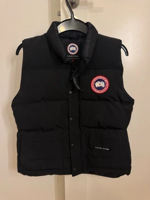Svart väst från Canada Goose - Svart dunväst från Canada Goose med logga på bröstet och krage. Storlek L / G. Kids size. Västen har två stora fickor framtill och stängs med knappar.  Materialet är en blandning av bomull och polyester med dunfyllning. Inga märken av användning.