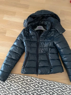 Blå pufferjacka  - Snygg blå pufferjacka från lager 157 i storlek XXS. Jackan har en huva, två dragkedjefickor. Materialet är glansigt och känns lätt, perfekt för kalla dagar. Dragkedja framtill och cool passform.