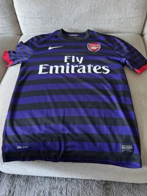 Svart och lila randig Arsenal Nike tröja - Arsenal matchtröja från Nike med svarta och lila horisontella ränder, röd detalj på ärmslut och vit logga. Fly Emirates tryck på bröstet och autenticitetsmärke nedtill. Tillverkad i Dri-FIT polyester som andas.