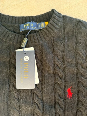Svart stickad tröja från Polo Ralph Lauren - Svart kabelstickad tröja från Polo Ralph Lauren med rund hals och röd broderad logga på bröstet. Tröjan har ribbade muddar vid hals, ärmslut och nederkant. Perfekt för dig som gillar klassisk och stilren design.