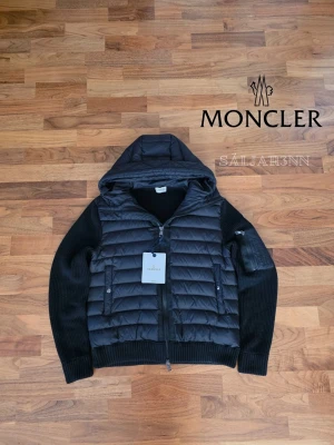 Svart pufferjacka från Moncler - Letar du efter den perfekta jackan inför våren? ☀️ Denna Moncler cardigan har quiltad dunfront och stickade ärmar för en stilren look. Observera att den är liten i storleken. Denna passar mer som M, trots att det står xl. Perfekt för både vardag och fest.   ✔ Lyxig design – Quiltad dun och stickat material. ✔ Praktisk – Huva, dragkedja och fickor. ✔ Ikonisk touch – Moncler-logga på ärmen.  Endast provad – nyskick! 📦Snabb leverans, Skickas samma dag📦