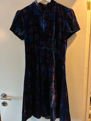 Mörkblå sammetklänning från Sandro - Supersnygg mörkblå sammetklänning från Sandro Paris med korta ärmar och färgstarkt paisleymönster i blått, lila och rött. Färgen skiftar beroende på ljus. Klänningen har hög hals, dragkedja bak och sitter fint i midjan. Lätt puffärm. Ca 90 cm lång. 