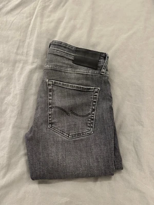 Grå jeans med svarta detaljer - Snygga grå jeans med svarta detaljer och klassisk femficksdesign. Bakfickan har en cool broderad våg och jeansen har en mörk tvätt som ger en schysst look. Passar perfekt till sneakers och hoodie. Materialet är mjukt och stretchigt jeans. Strl 30,30