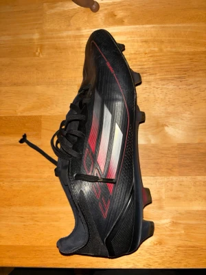 Svarta Adidas fotbollsskor med röd detalj - Säljer ett par svarta Adidas fotbollsskor med röda och silverfärgade detaljer. Skorna har klassisk snörning och de ikoniska tre ränderna på sidan. Yttermaterialet är syntet och sulan har dobbar för bra grepp på planen. Perfekt för dig som vill ha stil och funktion på träningen.