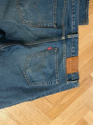 Levi's 514 blå jeans W32 L34 - Klassiska blå Levi's 514 jeans med raka ben och normal passform. Jeansen har fem fickor, bruna kontrastsömmar och det ikoniska läderpatchen bak i midjan. Tillverkade i slitstarkt denim av bomull. Perfekta för en avslappnad och tidlös stil.