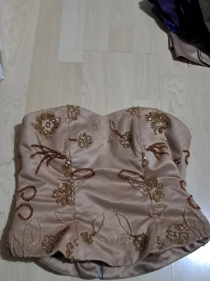 Champagnefärgad blommig korsett - Säljer en champagnefärgad korsett med blommiga broderier och paljettdetaljer i guld och brons. Toppen är axelbandslös och har en dragkedja bak för enkel påklädning. Materialet är glansigt och känns lyxigt, perfekt för att sticka ut. Storlek 38 eller en större S