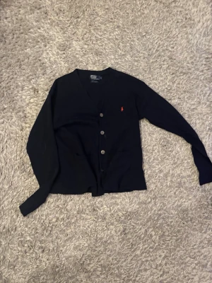 Mörk blå kofta från Polo Ralph Lauren - Mörk blå kofta från Polo Ralph Lauren med klassisk v-ringning och tre knappar framtill. Tröjan har långa ärmar, två fickor och den ikoniska röda Polo-loggan broderad på bröstet. Perfekt för lager-på-lager och stilrena outfits.