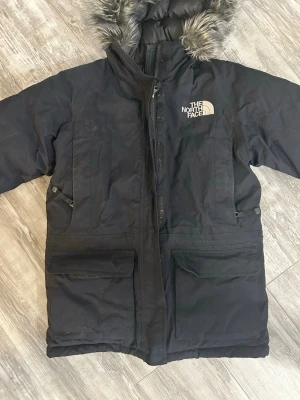 Svart dunjacka med fuskpäls från The North Face - Säljer en svart dunjacka från The North Face i storlek XS. Jackan har en fluffig huva med fuskpäls, stora fickor framtill och dragkedja. Perfekt för kalla dagar och har en schysst, klassisk look med broderad logga på bröstet.