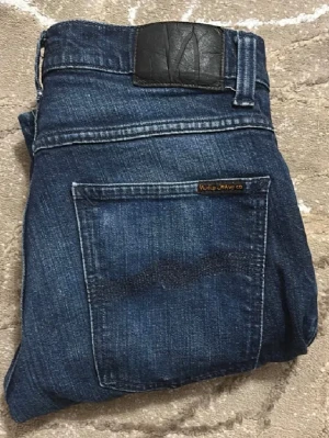 Mörkblå jeans från Nudie Jeans Co - Snygga mörkblå jeans från Nudie Jeans Co med klassisk femficksdesign och subtila slitningar. Jeansen har rak passform och är tillverkade i slitstarkt bomullsmaterial. Perfekt för dig som gillar stilrena och tidlösa jeans med coola detaljer.