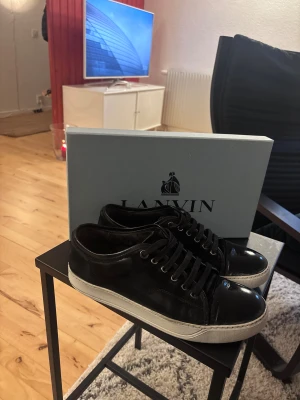 Svarta sneakers från Lanvin - Snygga svarta sneakers från Lanvin med vit platt sula och rund tå. Ovandelen är i mocka med lackad skinn-tå, och skorna har svarta snören och metallöglor. Perfekta för dig som gillar stilrena och lyxiga sneakers med exklusiv känsla. Målade med vit skofärg och använts i 2 år. Inga returer!
