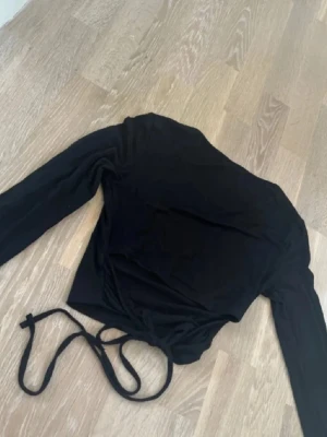 Svart långärmad omlottopp med snörning - Säljer en svart långärmad topp med knytning i ryggen. Toppen är tight och har en stilren look som passar till många olika outfits. Materialet känns mjukt och stretchigt. Perfekt för dig som gillar trendiga detaljer. Finns ett litet hål som lätt går att fixa.