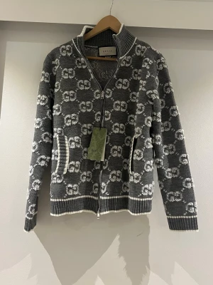 Grå stickad kofta från Gucci - Säljer en grå stickad kofta från Gucci med vit GG-logga som mönster över hela plagget. Koftan har dragkedja framtill, ribbade muddar och krage med vit kant. Perfekt för dig som gillar exklusiva märken och snygga detaljer.