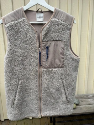 Beige teddyväst från Dobber, storlek S - Säljer en beige teddyväst från Dobber i storlek small. Västen har mjukt och fluffigt teddy-material, detaljer i beige och en bröstficka med dragkedja och blå snörning. Två sidofickor med dragkedja och en stilren look utan ärmar. Loosefit. Mer som medium. 