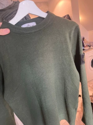 Grön Kashmir tröja från Stone Island - Snygg mörkgrön stickad tröja från Stone Island med klassisk rund hals och ribbade muddar. Tröjan har den ikoniska Stone Island-patchen på vänster ärm, vilket ger en extra cool detalj. Perfekt för dig som gillar streetwear och vill ha något stilrent.