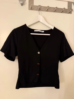 Svart blus med v-ringning från 157 - Säljer en svart blus från 157 i storlek S. Blusen har v-ringad hals, korta ärmar och tre dekorativa knappar framtill. Modellen är croppad och har en enkel, stilren design som passar till många olika looks.